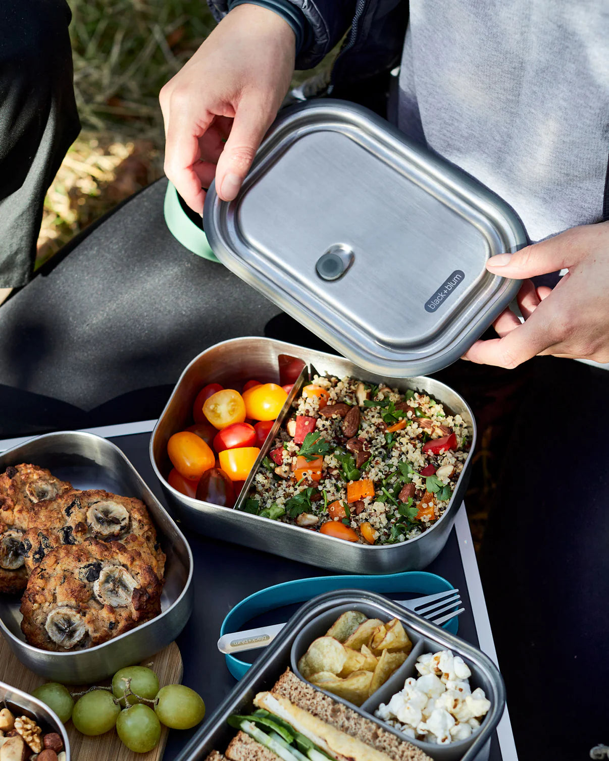 Person opening a metal lunch container with various food items on a picnic setup.