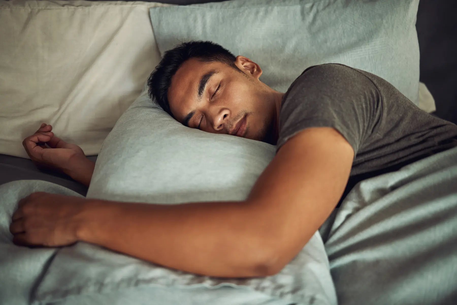 Body alignment during sleep illustrated by a man resting comfortably on his side hugging a pillow in soft gray bedding.