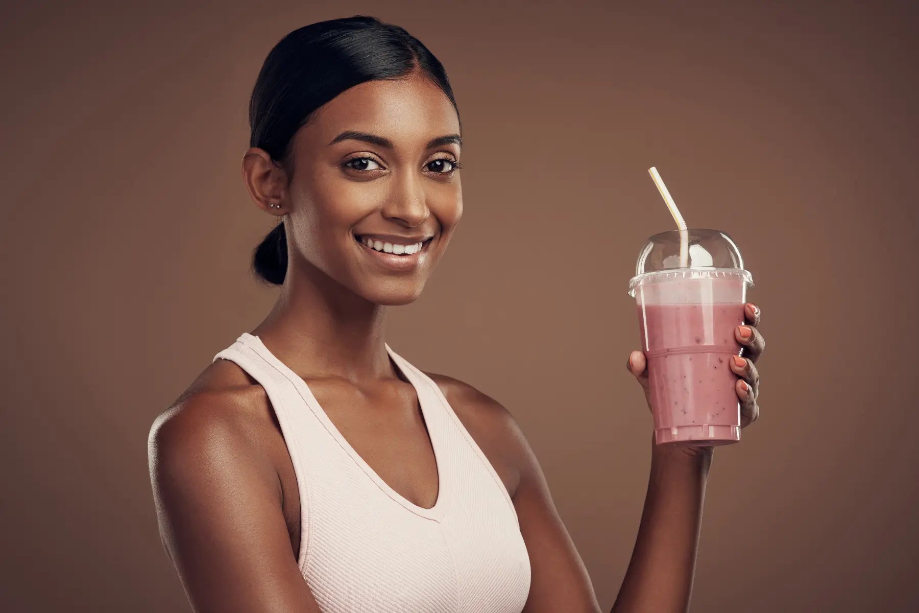 Body composition nutrition shown by a smiling woman in a sports bra holding a pink protein smoothie against a warm brown backdrop.