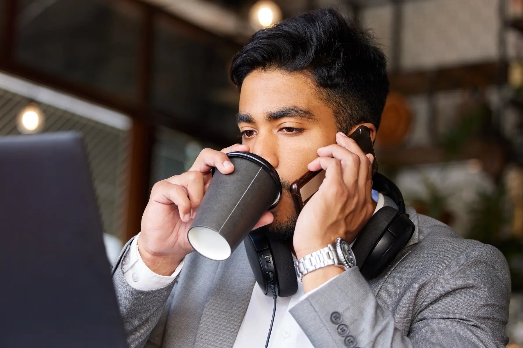 Desk life health impact busy man in a gray suit simultaneously drinking coffee and talking on the phone with headphones around his neck in front of a laptop.