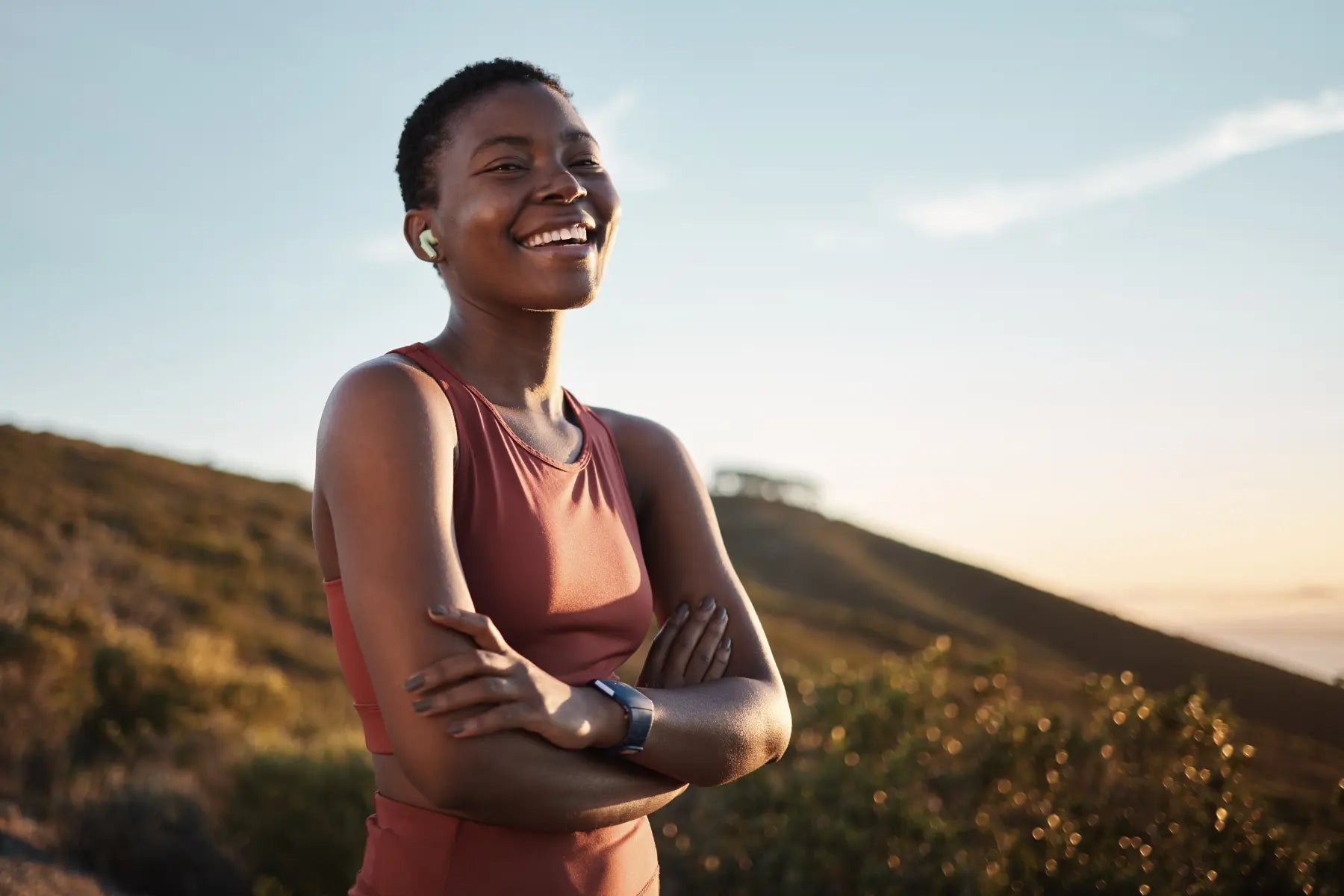 Fitness recovery success with a smiling woman in a red sports top standing arms crossed outdoors at golden hour with a fitness watch and earbuds.