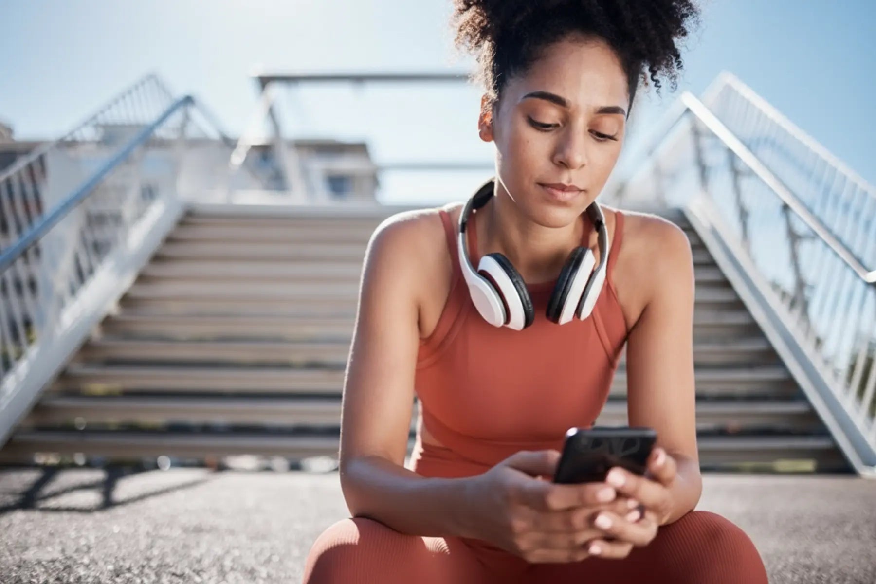 Performance tracking app checked by a young woman in orange activewear with headphones around her neck on outdoor steps.