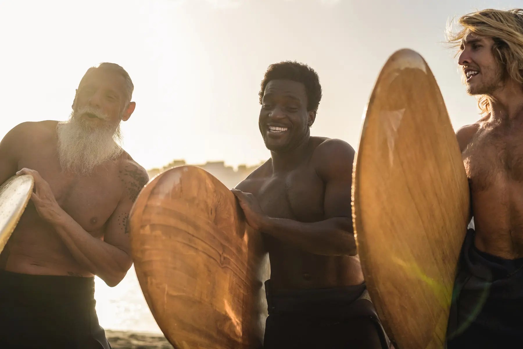Longevity and active aging shown by three smiling surfers of different generations holding wooden boards at golden hour on the beach.