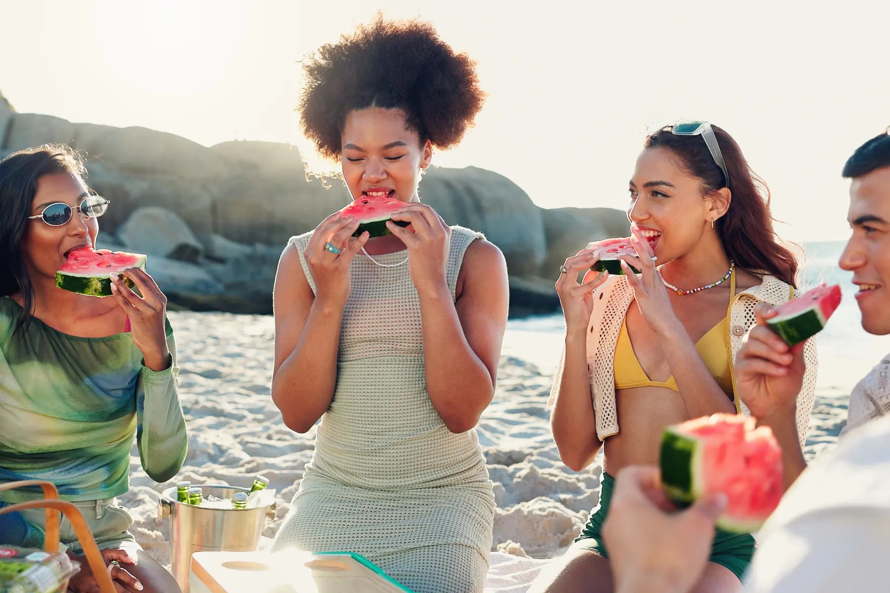 Summer eating habits with a group of friends biting into fresh watermelon slices on a sunny beach at golden hour.