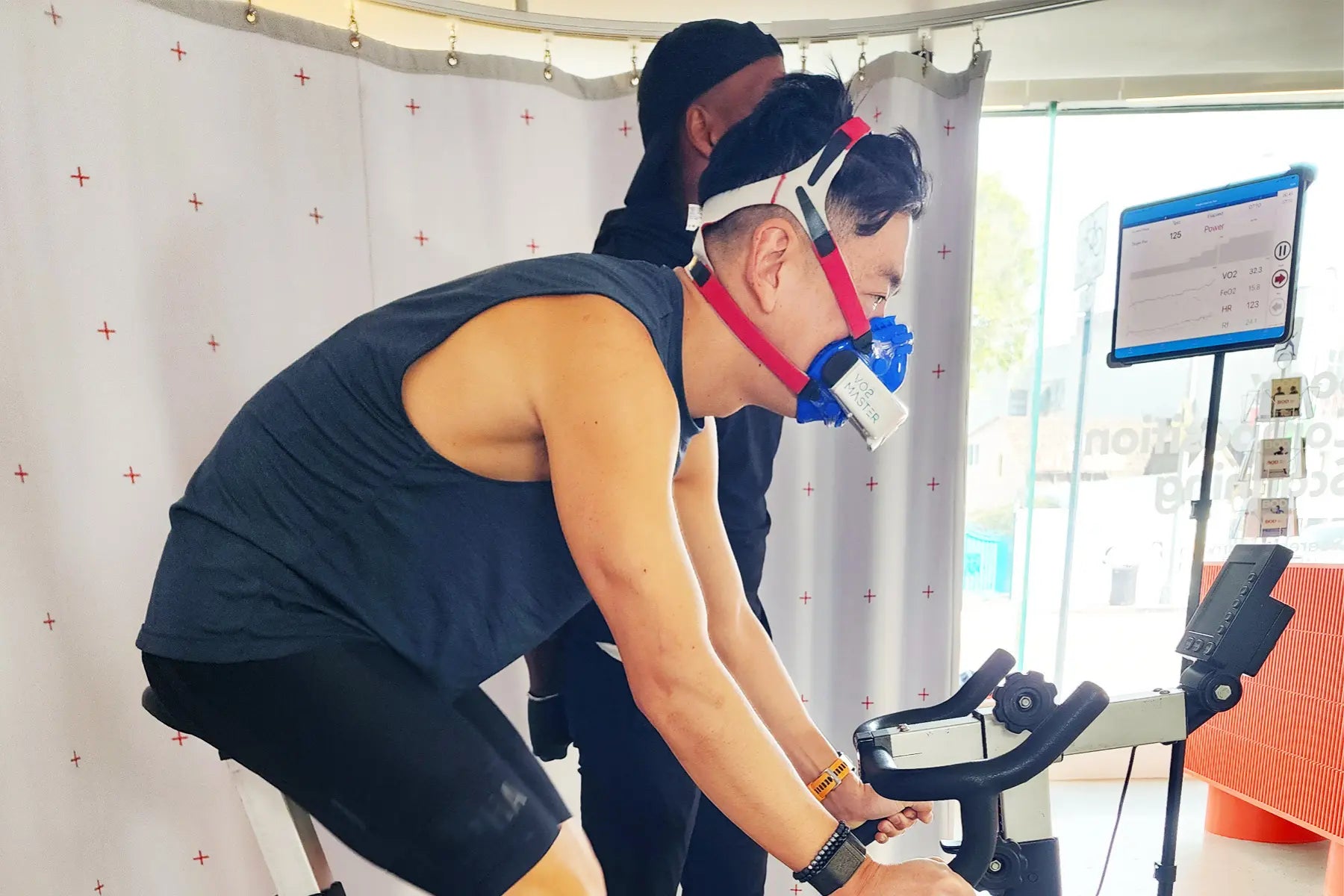 VO2 max test being performed by an athlete wearing a respiratory mask while cycling on a stationary bike connected to monitoring equipment in a lab setting.