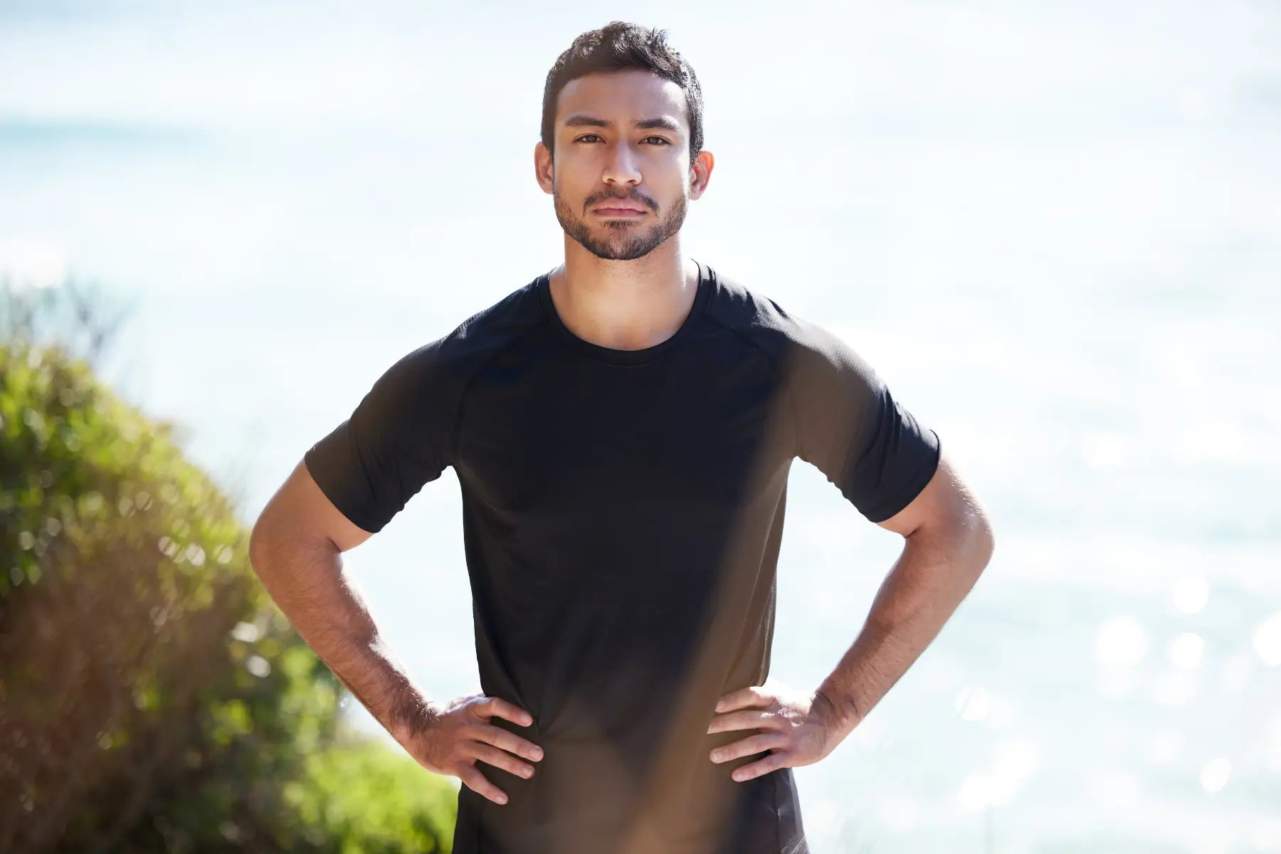 Visceral fat reduction goal with a fit man in a black t-shirt standing confidently with hands on hips against a bright coastal background.