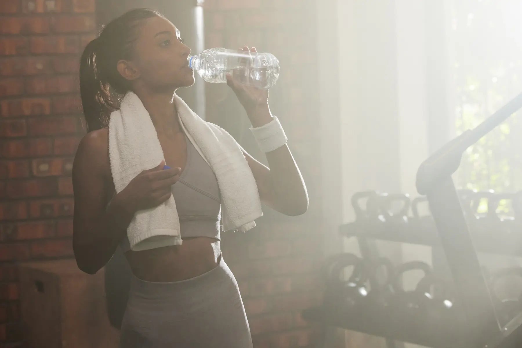 Aging and menopause during fitness training, woman in sports bra drinking water with a towel around her neck in a softly lit gym after a workout.