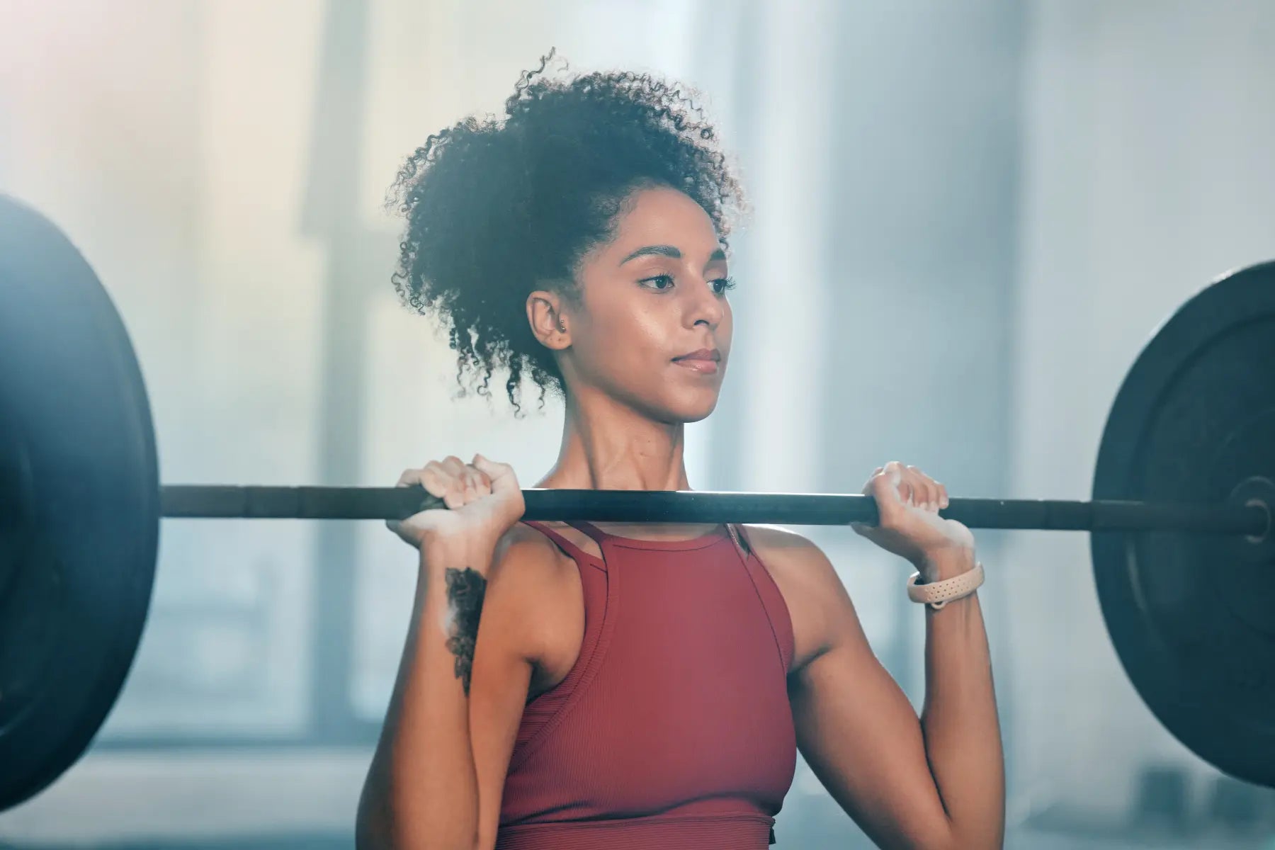 Bone density and health benefits of strength training shown as a focused young woman with curly hair back squats a loaded barbell in the gym.