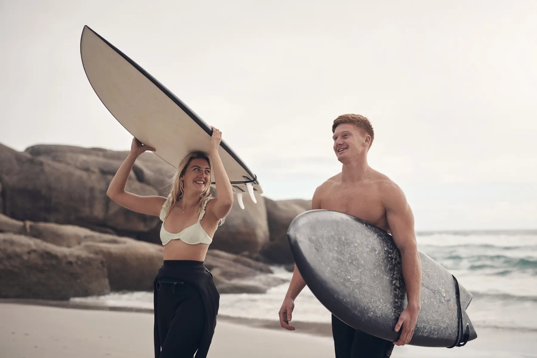 Bone health represented by fit surfers walking with surfboards on the beach near ocean rocks.