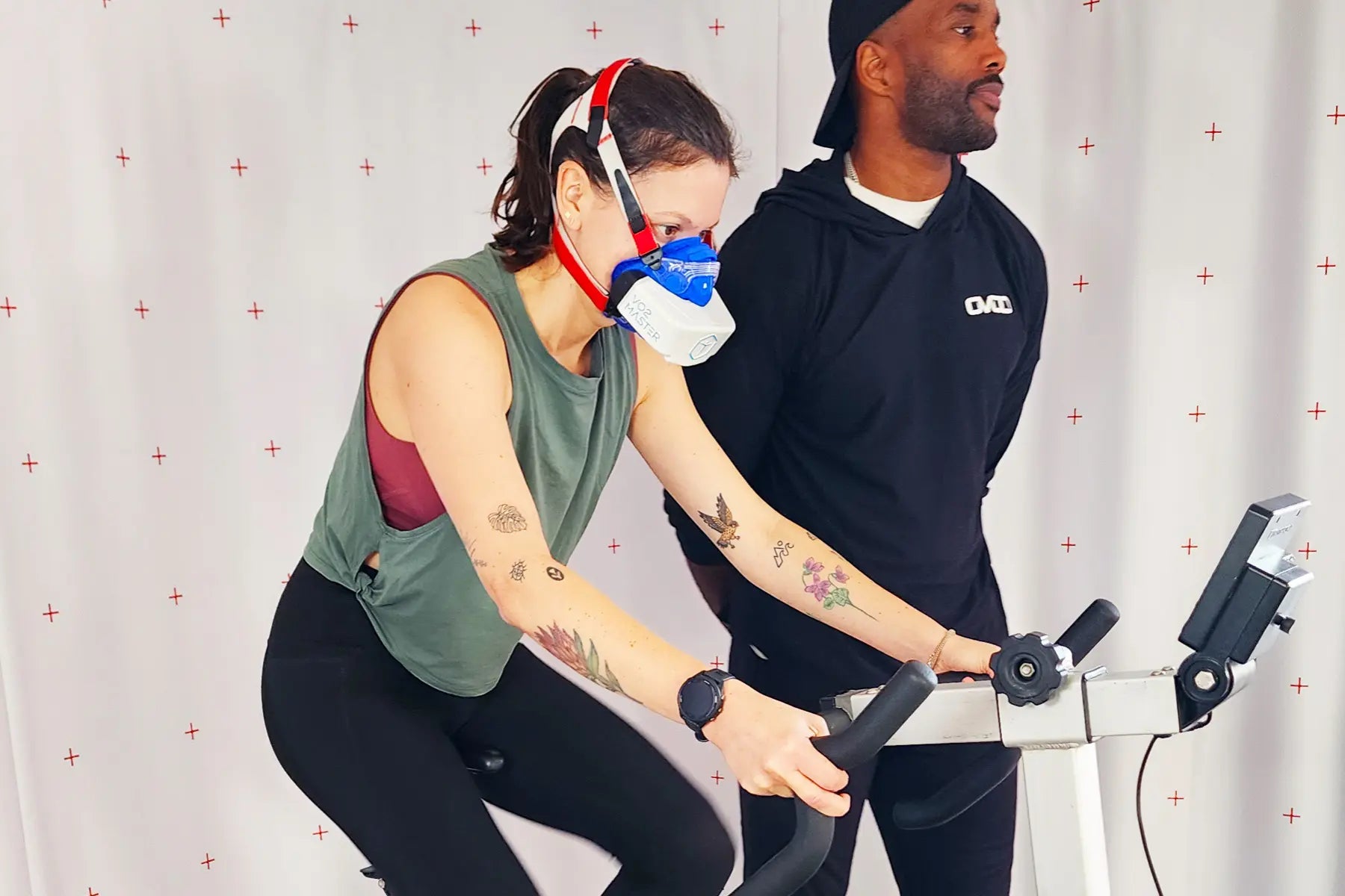 Cardiovascular endurance test in a lab setting with a woman cycling on a stationary bike while wearing a respiratory mask and monitored by a trainer.