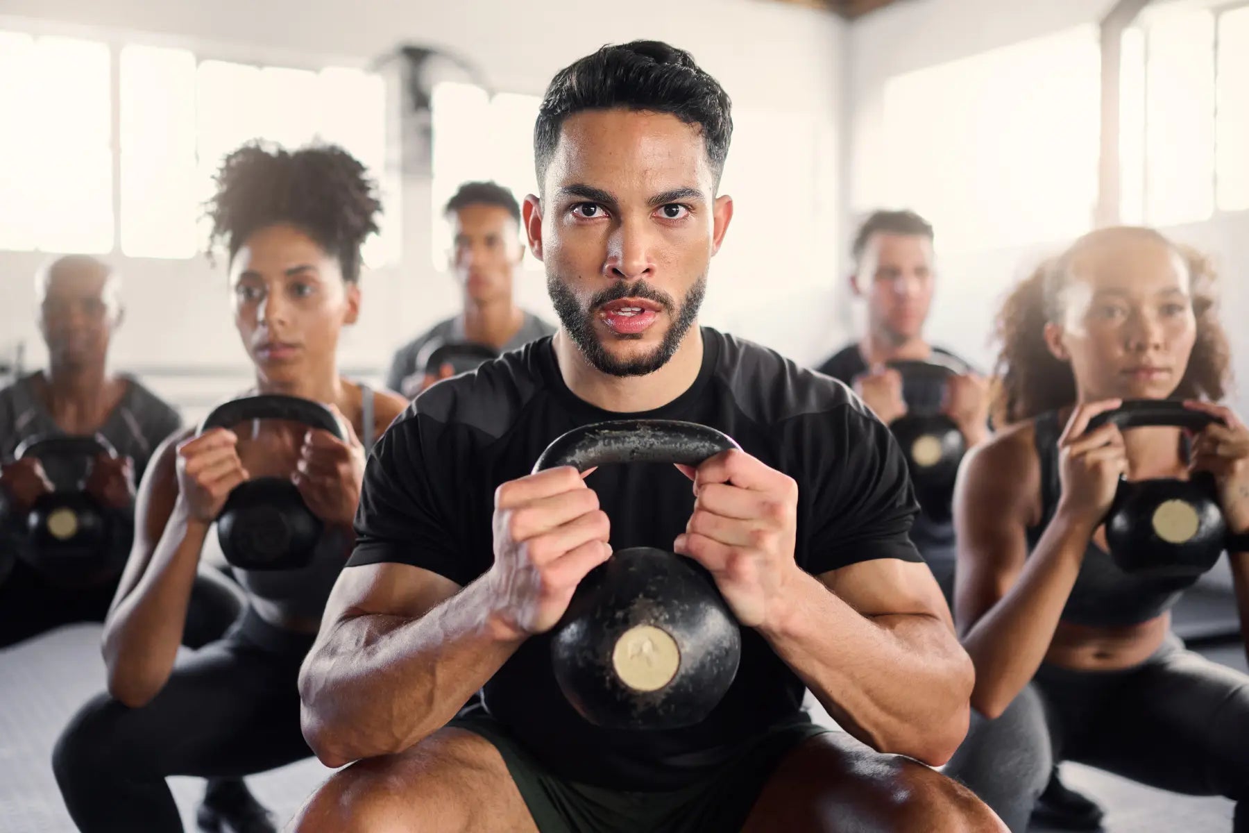 Fitness and fat levels improve through strength training, shown by group doing kettlebell squats in gym.