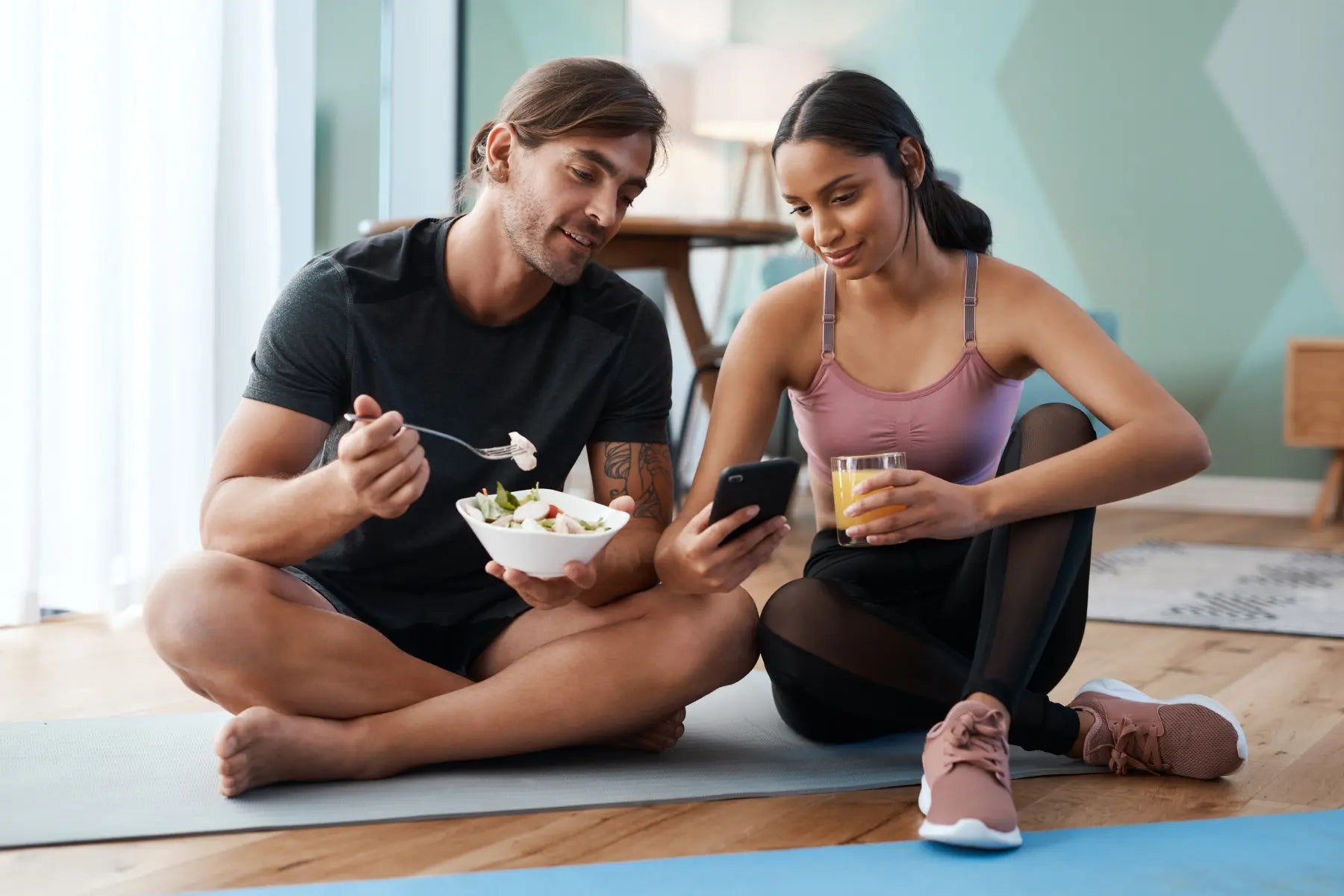 Healthy dietary fats featured in a post-workout meal as a couple sits on yoga mats sharing a salad and juice.