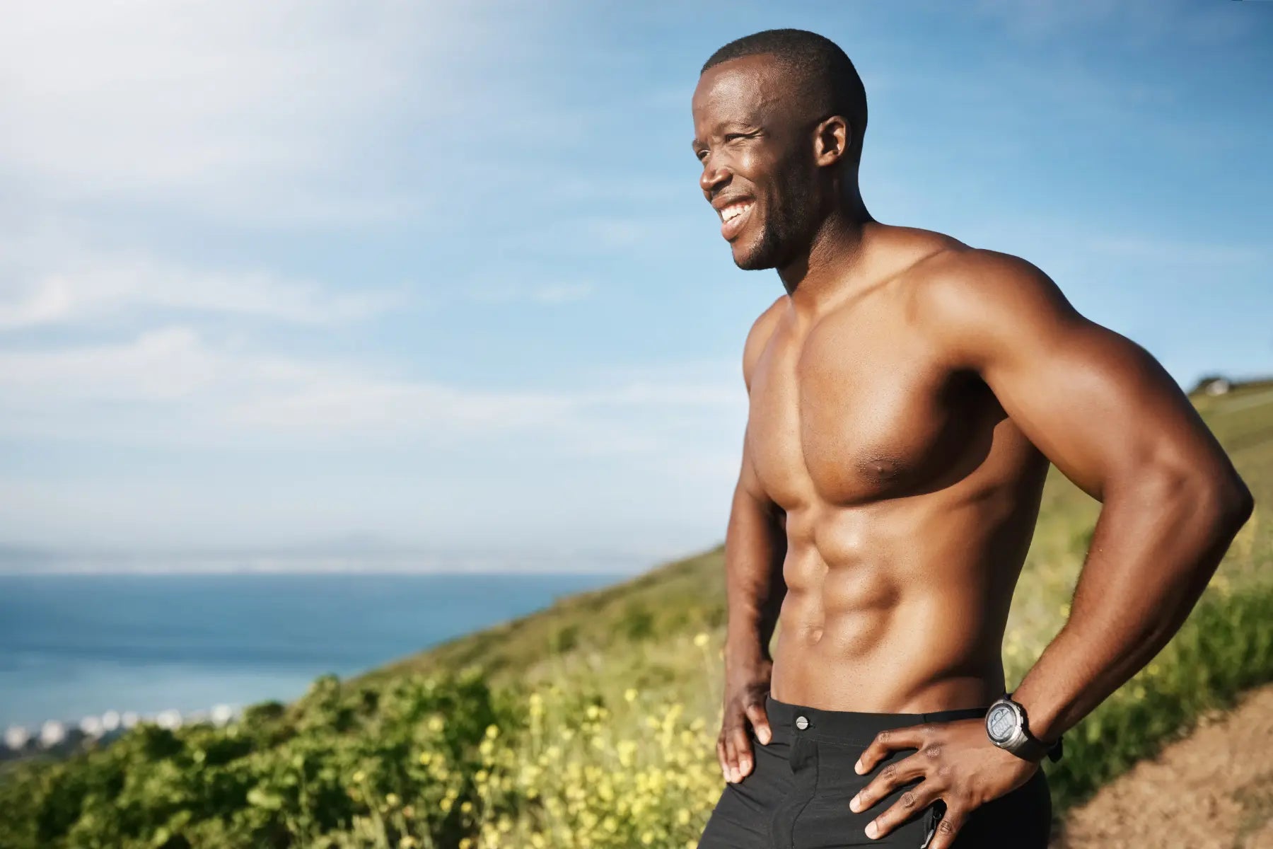 Muscle gain results visible on a lean, shirtless man with a defined six-pack smiling confidently on a scenic coastal hillside.