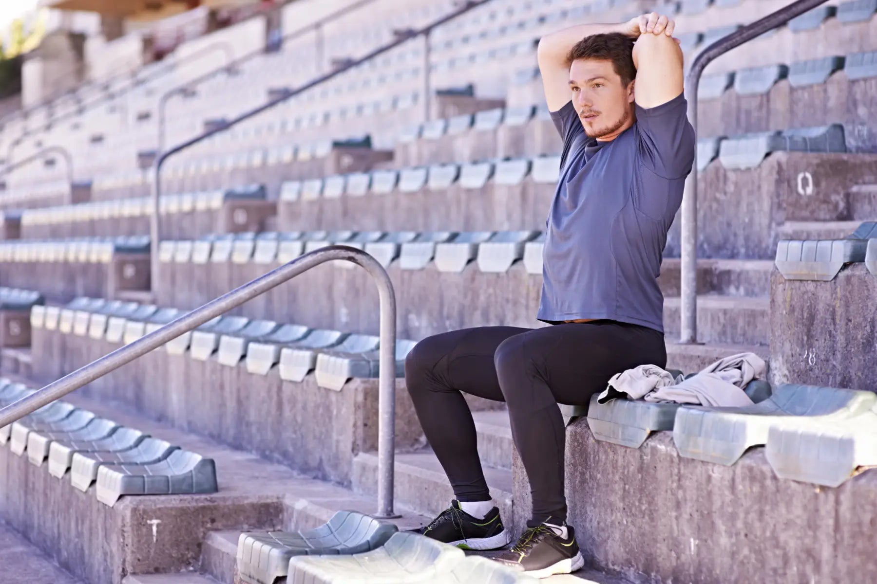 Post-workout stretching as a man extends his triceps while seated on empty stadium steps.