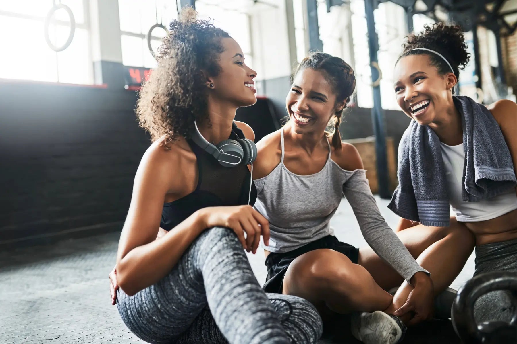 Three women in workout clothes laughing together while sitting on the gym floor after a workout.