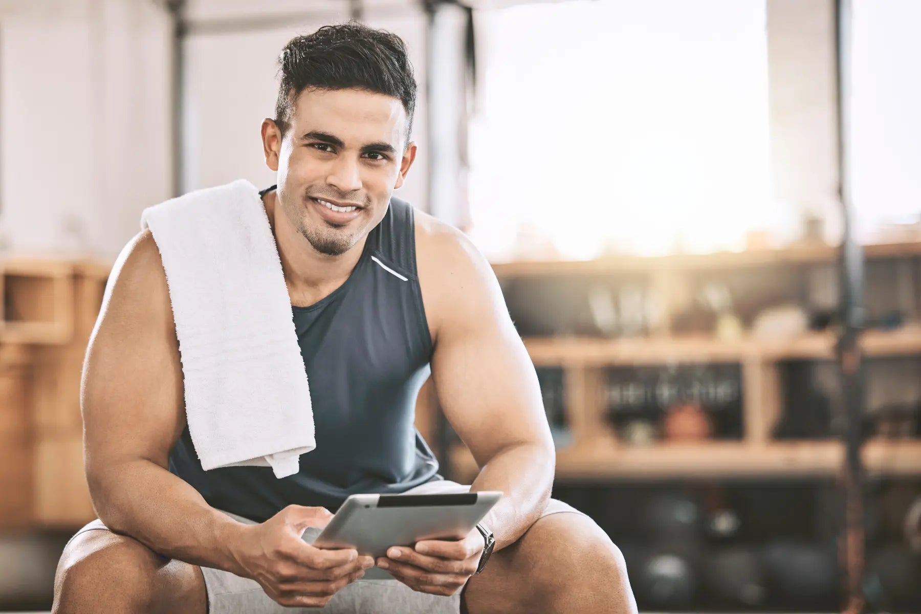 Testosterone replacement therapy consultation concept showing a muscular man with a towel over his shoulder sitting in a gym and holding a tablet.