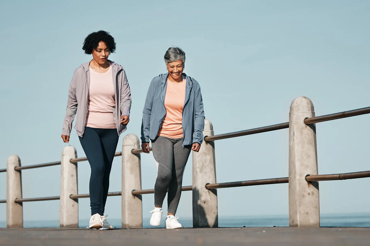 Two mature women in activewear walking along a seaside promenade, pairing exercise with weight loss medications.