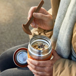 Person holding a insulated travel mug with a wooden spoon, sitting outdoors.