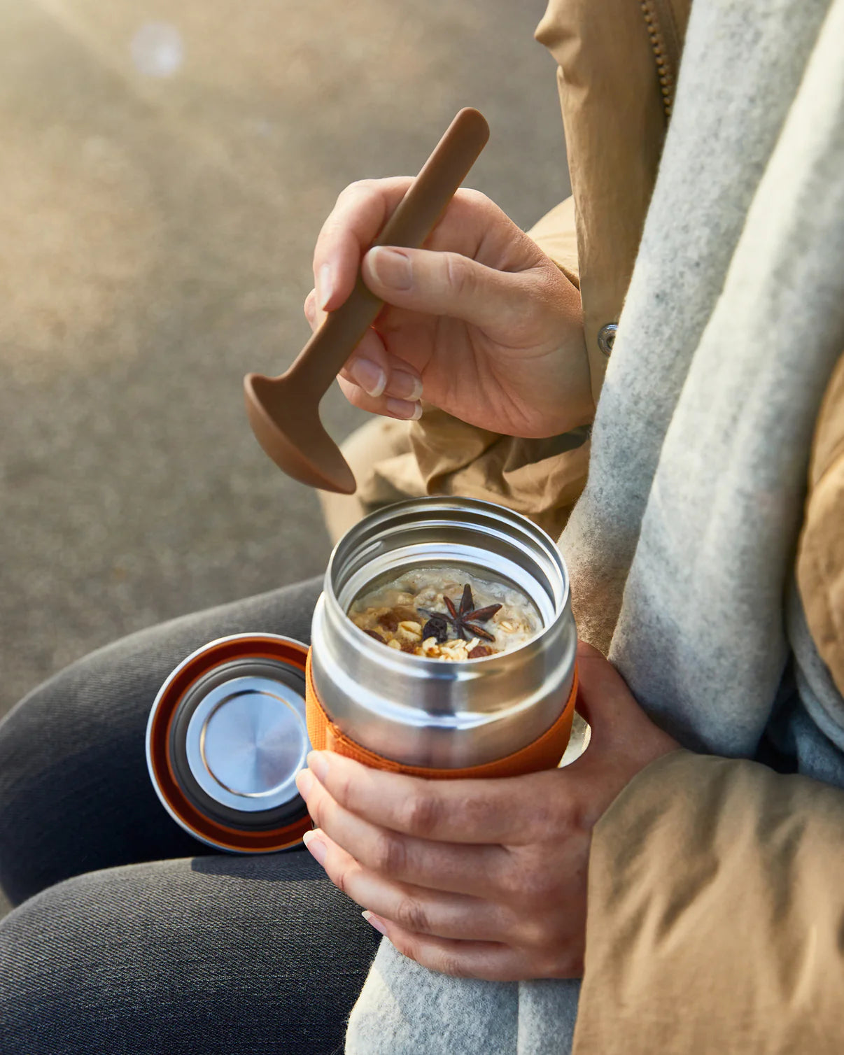 Person holding a insulated travel mug with a wooden spoon, sitting outdoors.