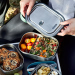 Person opening a metal lunch container with various food items on a picnic setup.